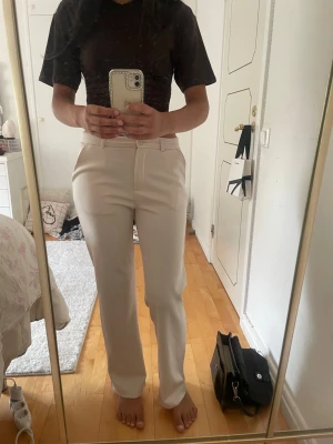 Beige kostymbyxa från 157 - Snygga beige kostymbyxor från 157 med raka ben och hög midja. Byxorna har bälteshällor, knapp och dragkedja framtill samt diskreta fickor. Perfekta för en clean och stilren look. Materialet känns mjukt och har en lätt struktur.dom är helt nya med lapp kvar