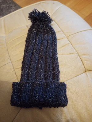 Svart  stickad mössa med tofs och glitter tråd stl one size  - Säljer en svart stickad mössa med glittertråd och stor tofs på toppen. Mössan har ribbad struktur och uppvikt kant, vilket ger en trendig och mysig look. Perfekt för kalla dagar när du vill sticka ut lite extra.
