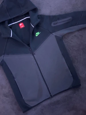 Svart och grå Nike Tech hoodie med dragkedja - Nike hoodie i svart och grått med hel dragkedja framtill och huva. Har en grön Nike-logga på bröstet och en snygg detalj på ärmen. Tillverkad i mjukt och bekvämt material, perfekt för en sportig och avslappnad stil. Skicket på hoodien är bra, inga synliga märken eller slitage. Priset är ej hugget i sten så går att diskutera! 