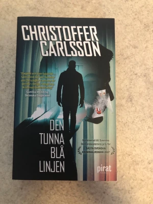 Den tunna blå linjen pocketbok - Spännande kriminalroman av Christoffer Carlsson, pocketutgåva från Piratförlaget. Omslaget går i blå och svarta toner med siluett av en person och stadsmiljö. Perfekt för dig som gillar deckare och svensk spänning.