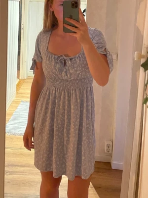 Ljusblå blommig klänning NA-KD - Supersöt ljusblå kortklänning från NA-KD x Pamela med små vita blommor, smock i midjan och knytband vid bysten. Puffiga kortärmar med knyt och en luftig, flowy kjol. Perfekt för sommarens alla tillfällen!