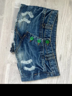 Kort jeanskjol med gröna knappar - Säljer en kort jeanskjol i blå denim med slitna detaljer och tre gröna knappar framtill. Kjolen har bälteshällor och synliga sömmar i kontrastfärg. Perfekt för dig som gillar en edgy och avslappnad stil.