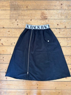 BACK år 2015; Svart kjol med logotypresår och dragkedja fram storlek 34 - BACK år 2015; Svart kjol med bred logotypresår. Kjolen har en dragkedja framtill och en ficka med knappdetalj. Materialet är lyocell. Perfekt skick! Storlek 34