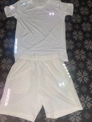 Vitt träningsset från Under Armour - Säljer ett vitt träningsset från Under Armour med t-shirt och shorts. Setet har reflekterande loggor och textdetaljer, samt elastisk midja med dragsko på shortsen. Perfekt för gymmet eller löpning. Materialet är lätt och andas bra.