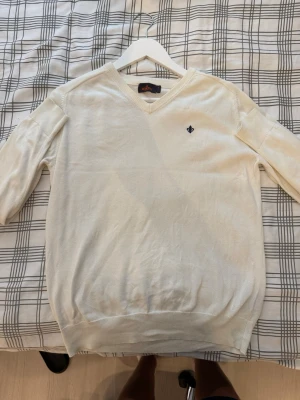 Vit stickad Morris tröja med V-ringning - Vit stickad tröja med V-ringning och diskret broderad logga på bröstet. Tröjan har långa ärmar och ribbade muddar vid ärmslut och nederkant. Perfekt för en stilren och enkel look.