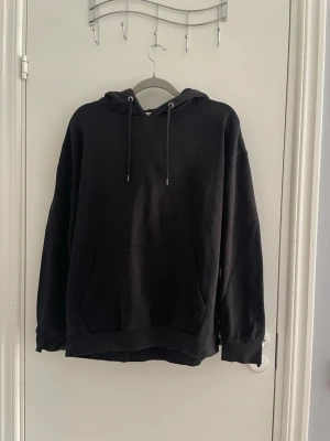 Svart hoodie med magficka - En klassisk svart hoodie med huva och dragsnören. Hoodien har en stor magficka framtill och ribbade muddar vid ärmslut och nederkant. Perfekt för en avslappnad och stilren look. Materialet ser ut att vara mjukt och bekvämt, troligen bomullsblandning.