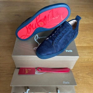 Marinblå sneakers från Louboutin - Snygga marinblå sneakers i mocka från Christian Louboutin med ikonisk röd sula och diskret logga undertill. Skorna har klassisk låg profil och ton-i-ton snörning för en clean look. Perfekta för dig som vill sticka ut med exklusiv stil.