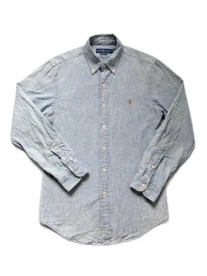 Blå skjorta från Ralph Lauren - Klassisk ljusblå skjorta från Ralph Lauren med button-down krage och broderad logga på bröstet. Tillverkad i mjuk bomull och har långa ärmar samt knappar framtill. Perfekt för en clean och stilren look.