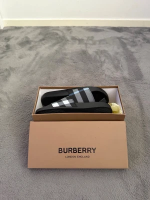 Burberry rutiga slides svart/vit - Snygga slides från Burberry i svart med klassiskt rutigt band i svart, vitt och grått. Tillverkade i slitstark gummi med räfflad sula för bra grepp. Perfekta för en avslappnad och trendig look.