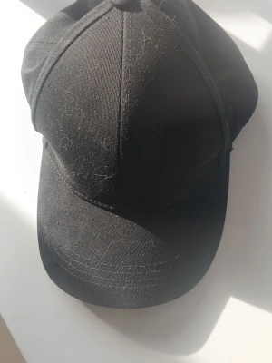 Svart klassisk keps - En stilren svart keps med böjd skärm och sex paneler. Kepsen är tillverkad i ett mjukt tygmaterial och har en enkel, tidlös design utan synligt märke eller dekorationer. Perfekt för en avslappnad streetwear-look.