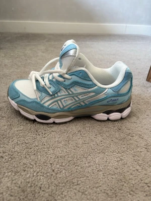 ASICS GEL blåvita sneakers - Snygga sneakers från Asics i ljusblått och vitt med lila logga på plösen. Ovandelen är i mesh och mockaliknande material, med vita skosnören och detaljer i grått. Skorna har en chunky sula och sportig vibe, perfekta för en trendig streetstyle.