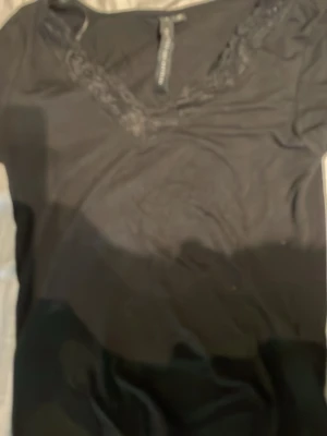 Svart topp med spetsdetaljer - Säljer en svart topp med v-ringad hals och snygga spetsdetaljer längs ringningen. T-shirten har korta ärmar och är gjord i ett mjukt material som känns skönt mot huden. Perfekt att matcha med jeans eller kjol.