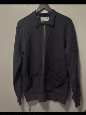Mörkgrå ziptröja från Selected Homme - Mörkgrå långärmad ziptröja från Selected Homme med krage och två framfickor. Tröjan har ribbade muddar vid ärmslut och nederkant, perfekt för lager-på-lager. Enkel och stilren design som passar till det mesta.