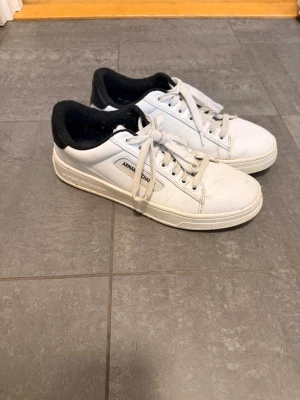 Vita sneakers från Armani Exchange - Snygga vita sneakers från Armani Exchange med svart vadderad häl och vita snören. Klassisk låg modell med diskret logga på sidan och gummisula. Perfekta för en clean och stilren look. Varm insida 