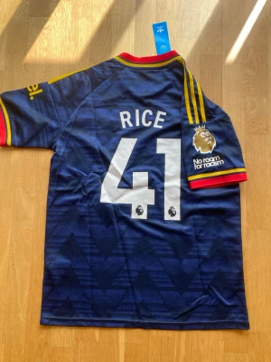Arsenal matchtröja Rice #41 blå 26/27 away - Snygg Arsenal matchtröja i mörkblått med gula detaljer och röda inslag vid krage och ärmslut. Adidas-logga och klubbmärke framtill, samt 'Emirates Fly Better' tryck. Nummer 41 och namnet Rice på ryggen. Premier League-märke på ärmen.