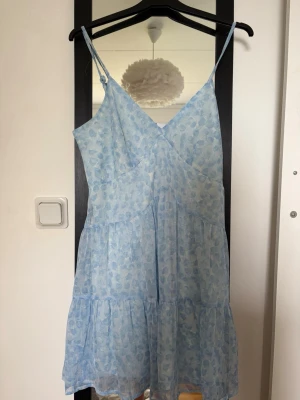 Ljusblå blommig sommarklänning - Söt ljusblå klänning med smala axelband och v-ringning. Klänningen har ett blommigt mönster, volangdetaljer och är tillverkad i ett lätt, luftigt material. Perfekt för varma dagar och ger en romantisk känsla.
