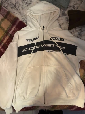Vit Corvette hoodie med dragkedja - Säljer en vit hoodie med dragkedja och tryck framtill med texten 'Corvette Grand Sport', racingdetaljer och fickor. Hoodien har huva med dragsko och ribbade muddar. Perfekt för dig som gillar bilar och sportig stil.