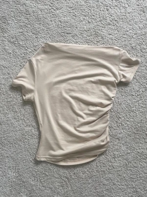 Beige asymetrisk topp - Superfin beige asymmetrisk topp. Sitter offshoulder och scruchat på ena sidan. Fått i present men klippt av lapp, vet därför inte märket. Perfekt basplagg till sommaren eller som intressant topp för layering under hösten. Knappt använd. Skriv vid intresse!!💞