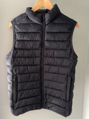 Marinblå väst  - Snygg marinblå dunväst med quiltad design och krage. Västen har dragkedja framtill och två sidofickor. Perfekt att slänga över en hoodie eller tröja för extra värme. Materialet är lätt och smidigt, vilket gör den enkel att bära med sig.