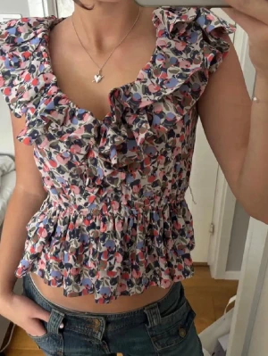 Blommig volangblus med v-ringning - Supersöt blommig blus med volanger och v-ringning. Toppen har ett färgglatt mönster i rosa, blått och grönt på vit botten, är ärmlös och har smock i midjan för en feminin passform. Perfekt för dig som gillar romantisk stil och detaljer.