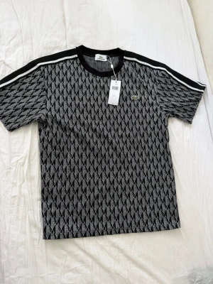 Svart mönstrad t-shirt från Lacoste - Säljer en svart t-shirt från Lacoste med vitt geometriskt mönster och klassisk krokodillogga på bröstet. T-shirten har svarta och vita ränder på axlarna, rund halsringning och är tillverkad i mjuk bomull. Perfekt för dig som gillar sportig och stilren design.