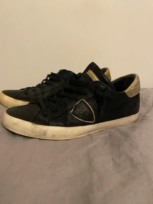 Svarta sneakers med gulddetaljer - Snygga svarta sneakers med guldfärgade detaljer på häl och plös samt en guldkantad patch på sidan. Skorna har vita sulor och svarta skosnören. Materialet ser ut att vara mocka eller liknande textur. Perfekta för dig som gillar en stilren men ändå unik look. Skicka gärna bud