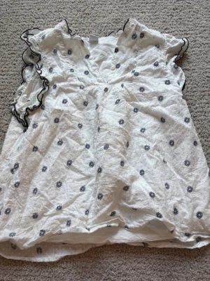 Vit blus med svarta broderade blommor - Söt vit blus med svarta broderade blommor och volangdetaljer vid ärmarna. Blusen har v-ringning och korta ärmar med svart kant. Perfekt för en trendig och somrig look. Materialet känns lätt och luftigt.