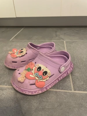 Lila tofflor med söta figurer - Säljer ett par barn lila tofflor i plast med söta figurer och jordgubbe framtill. Tofflorna har texten 'Wish you Life is getting better and b...' längs sidan och justerbar hälrem. Perfekta för dig som gillar gulliga detaljer och bekväma skor.