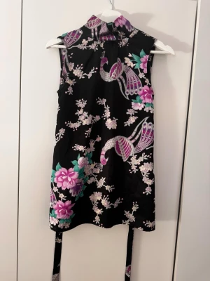 3 blommiga toppar, Svart topp med blommor, lila topp m blom, blå… - Snygg svart kort klänning med asiatiskt inspirerat blommönster i rosa, lila och grönt samt fågelmotiv. Klänningen är ärmlös, har hög krage och knytband i midjan. Perfekt för dig som vill sticka ut med en unik och färgglad look.