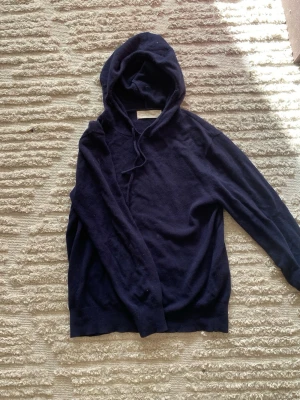 Mörkblå cashmere hoodie Sayles - Mysig mörkblå hoodie från Sayles i mjuk cashmere. Klassisk modell med huva och dragsko, samt långa ärmar och ribbade muddar. Perfekt för dig som vill ha en stilren och bekväm hoodie i ett lyxigt material.