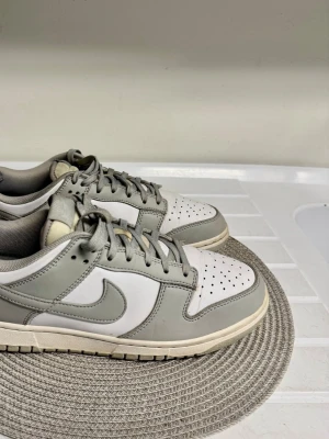 Nike Dunk Low gråvita sneakers - Nike Dunk Low sneakers i en clean grå och vit färgkombination. Skorna har klassisk låg silhuett, perforerad tåbox och ikoniska swoosh-loggan på sidan. Ovandelen är i läder med matchande grå snören och vit sula. Perfekta för dig som gillar stilrena och tidlösa sneakers.
