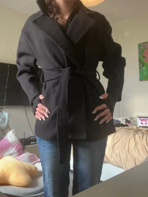 Svart kappa med knytskärp - Stilren svart kappa med bred krage och knytskärp i midjan. Kappan har en rak passform och är tillverkad i ett tjockare tyg som ger en snygg siluett. Perfekt för dig som vill ha en klassisk och enkel look.