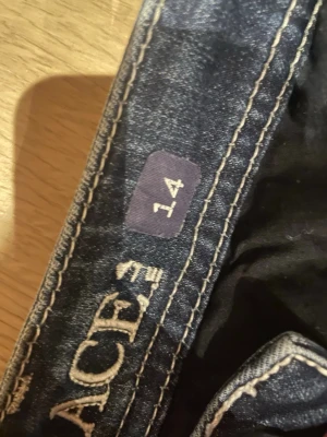 Mörkblå bootcut jeans stl 14 - Säljer ett par mörkblå bootcut jeans med snygga slitningar och kontrastsömmar. Jeansen har klassisk femficksmodell och är tillverkade i ett mjukt denimtyg som sitter skönt. Perfekta för dig som gillar en avslappnad men trendig look.