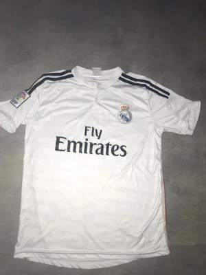 Real Madrid Ronaldo matchtröja - Säljer en vit Real Madrid matchtröja med svarta detaljer och tryck. Tröjan har klubbmärke och Fly Emirates-logga fram, samt Ronaldo och nummer 7 på ryggen. Korta ärmar och La Liga-märke. Perfekt för fotbollsfans!
