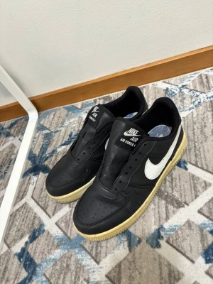 Nike Air Force 1 svarta sneakers - Säljer ett par klassiska Nike Air Force 1 sneakers i svart läder med vit swoosh och beige sula. Skorna har lågt skaft och perforerad tå för extra ventilation. Perfekta för dig som gillar streetwear och tidlös design.