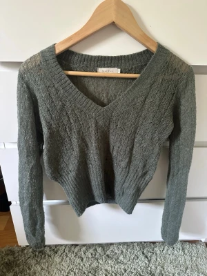 Grågrön stickad tröja med v-ringning - En stickad tröja i grönt med v-ringning och långa ärmar. Tröjan har ribbstickad nederkant och är tillverkad i ett mjukt, tunt material som ger en avslappnad look. Perfekt att matcha med jeans eller kjol.