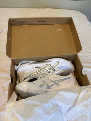 Vita Asics  - Vita Asics med silvriga detaljer på sidorna och meshpaneler för extra andningsförmåga. Perfekt för innomhusbruk av alla olika aktiviteter. 