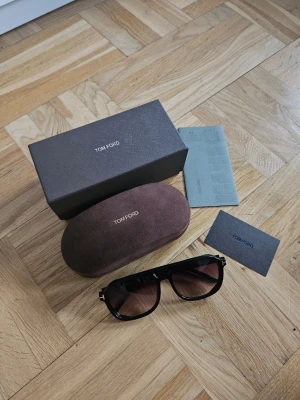 Tom Ford Rosco pilotbågar solglasögon - Snygga svarta solglasögon från Tom Ford med klassisk pilotform, modellnamn Rosco. Glasen är tonade i faded mörkbrunt och bågarna har en stilren design med diskreta gulddetaljer vid gångjärnen. Kommer med originalfodral och låda.