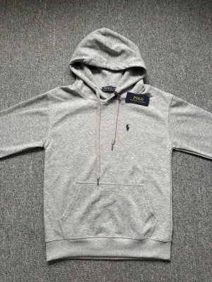 Grå hoodie från Polo Ralph Lauren - Snygg grå hoodie från Polo Ralph Lauren med klassisk huva och dragsnören. Har en broderad logga på bröstet och en stor magficka framtill. Tillverkad i mjukt material som känns skönt mot huden. Perfekt för en avslappnad och stilren look. Har dock några defekter.