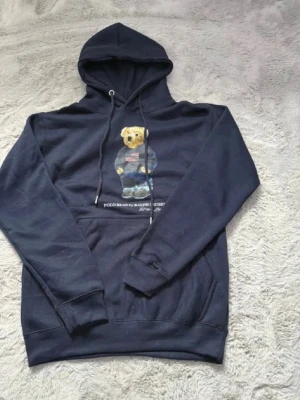 Polo Bear hoodie från Ralph Lauren - Två hoodies från Ralph Lauren med den ikoniska Polo Bear-trycket på bröstet. En är marinblå och den andra vit, båda med huva och känguruficka. Klassisk streetwear-stil med dragsko i huvan och mjukt material, perfekt för en avslappnad look.