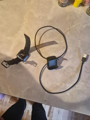 Svart smartklocka med laddare, Fitbit versa 2 - Säljer en svart smartklocka med fyrkantig display och silikonarmband. Klockan har touchskärm och kommer med en USB-laddare. Perfekt för att hålla koll på tid, steg och notiser direkt på handleden.