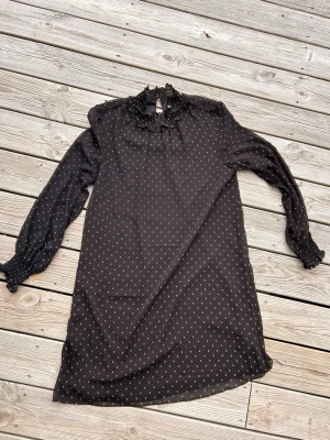 Svart prickig långklänning med volang - Svart långklänning med små vita prickar, långärmad och med volangdetaljer vid halsringningen. Klänningen har en hög krage med rynk och volang, samt smock vid ärmsluten. Perfekt för dig som gillar en romantisk och trendig stil. Tunna ärmar. Knälång