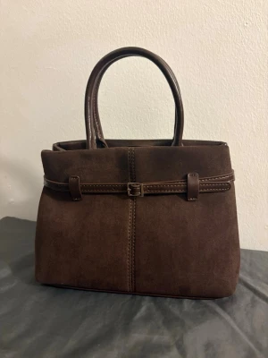 Brun handväska med bältdetalj - Snygg handväska i mörkbrun mocka med matchande handtag och en dekorativ bältdetalj runt väskan. Baksidan är i slätt brunt skinnliknande material. Perfekt storlek för vardagens essentials och ger en stilren look.