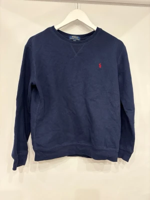 Mörkblå sweatshirt från Polo Ralph Lauren - Klassisk mörkblå sweatshirt från Polo Ralph Lauren med rund halsringning och den ikoniska röda logotypen broderad på bröstet. Tröjan har ribbade muddar vid ärmslut och nederkant, perfekt för en avslappnad och stilren look.