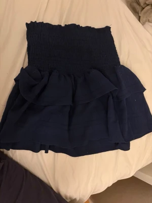 Mörkblå volangkjol med smock - Supersöt mörkblå kortkjol med smock i midjan och volangdetaljer. Kjolen har en stretchig passform och är gjord i ett mjukt bomullsliknande material. Perfekt att styla med en enkel topp eller hoodie för en chill look.