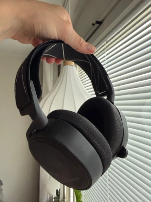 Svarta SteelSeries Arctis 9  trådlösa hörlurar - Säljer ett par svarta over-ear hörlurar från SteelSeries med mjuka tygkuddar och justerbart huvudband med mönster. Trådlös modell med tillhörande USB-sändare och laddkabel. Perfekta för gaming eller musik med bekväm passform och stilren design.