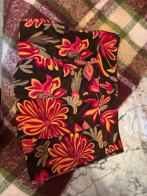 Brun blommig minikjol med färgglada mönster - Säljer en brun minikjol med stora blommiga mönster i rött, orange och gult. Kjolen har en rak passform och är sydd i ett mjukt tyg. Perfekt för dig som gillar färgstarka plagg och vill sticka ut lite extra. Med en liten dragkedja på ena sidan 