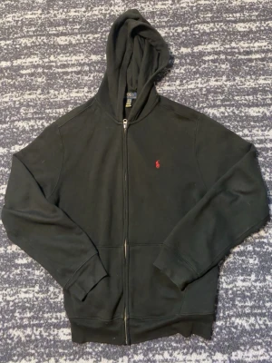 Svart hoodie med dragkedja från Polo Ralph Lauren - Säljer en svart hoodie med dragkedja från Polo Ralph Lauren. Klassisk design med huva, ribbade muddar och fickor framtill. Liten röd logga broderad på bröstet. Perfekt för en avslappnad och stilren look. XL i barnstorlek så den är S eller XS för äldre.