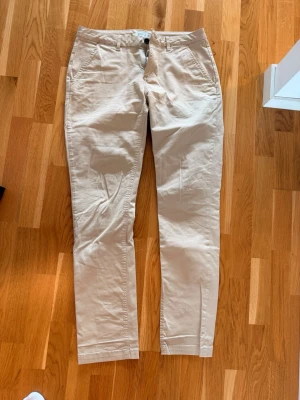 Beige chinos från Mouli - Snygga beige chinos från Mouli i storlek 32. Klassisk rak passform med fickor fram och bak, samt bälteshällor i midjan. Tillverkade i mjukt och bekvämt bomullsmaterial som passar perfekt till en avslappnad stil.