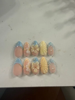 Blå och gula press-on naglar med 3D-blommor - Fina nagelband för sommaren. Naglarna är gjorda med uppbyggande geler så att de håller länge utan att gå sönder. Nagelstorlekar från tumme till lillfinger: 17 mm, 13 mm, 15 mm, 14 mm, 11 mm.  Inkluderar fäste: nagellim, nagelstickers, nagelfil, buffert och nagelbandspress. 